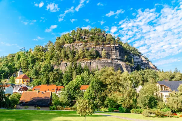 Blick auf einen Berg mit großen sichtbaren Felsabschnitten und Bäumen vor blauem, leicht bewölktem Himmel. Im Vordergrund sind eine Wiese und einzelne Häuser einer Ortschaft zu sehen.