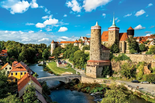 Ein Fluss mit einer kleinen Brücke. Rechts von ihr ist ein alter Stadtturm mit Stadtmauer und weiteren alten Gebäuden zu sehen.
