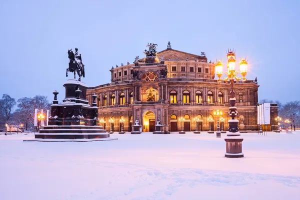 Die beleuchtete Semperoper in Dresden im Winter mit Schnee am Vorplatz und auf der Statue, die sich darauf befindet.
