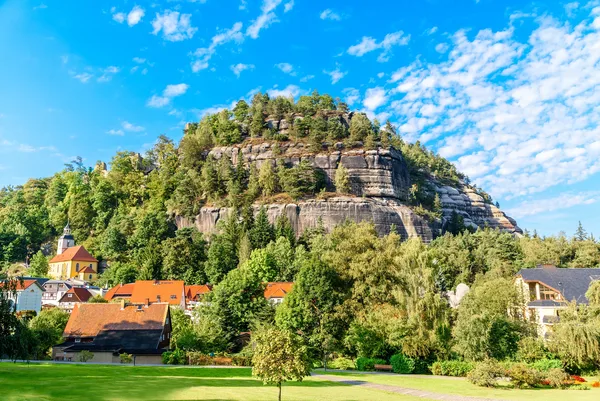Blick auf einen Berg mit großen sichtbaren Felsabschnitten und Bäumen vor blauem, leicht bewölktem Himmel. Im Vordergrund sind eine Wiese und einzelne Häuser einer Ortschaft zu sehen.