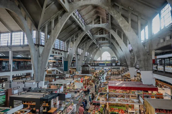 Eine Halle mit hoher Decke und Bögen aus Beton, in der viele Stände mit Obst, Gemüse und anderen Lebensmitteln aneinander gereiht stehen