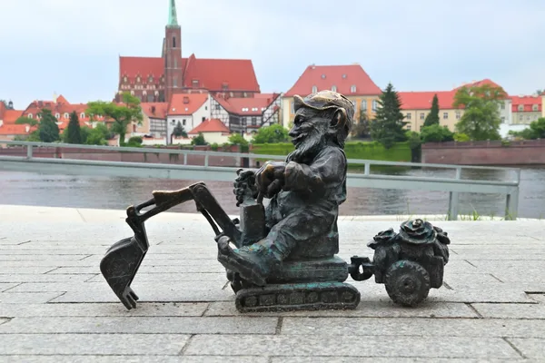 Eine kleine Zwergenstatue aus Eisen, die einen Bagger mit Anhänger bedient. Im Hintergrund ist eine Fluss und eine Stadt zu sehen.