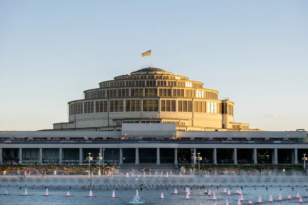 Blick auf die große Jahrhunderthalle in Wrocław mit hoher mehrstöckiger Kuppel im oberen Bereich und einer großen Wasserfläche mit einzelnen kleinen Fontänen im Vordergrund