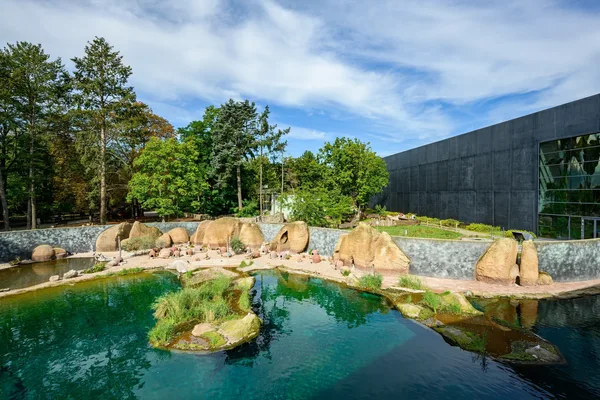 Ein künstlich angelegter See mit kleinen bepflanzten Inseln und einem kleinen Strandbereich, der von einer niedrigen Mauer eingezäunt ist. Im Hintergrund sind Bäume und ein großes Gebäude mit dunkler Fassade zu sehen.