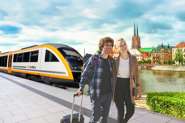 Ein Paar aus Mann und Frau spaziert mit einem Reisetrolley auf dem Bahnsteig weg vom trilex. Im Hintergrund ist Wrocław zu sehen.