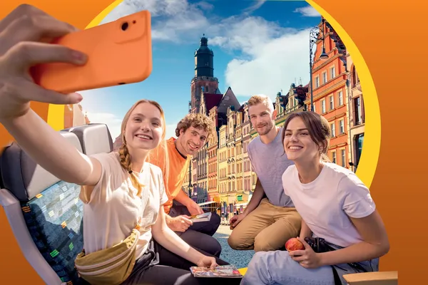 Zwei junge Frauen und zwei junge Männer sitzen auf Zugsitzen und machen ein Selfie. Im Hintergrund ist in einem runden Bildausschnitt eine bunte Häuserfassade zu sehen.