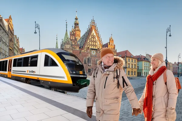 Ein Paar aus Mann und Frau spaziert in Winterklamotten im Vordergrund des Bilds. Im Hintergrund sind der trilex und Häuser in Wroclaw zu sehen.