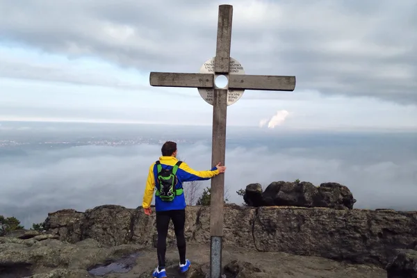 Mann steht in Wanderkleidung mir dem Rücken zum Betrachtenden an einem Gipfelkreuz und berührt dieses.