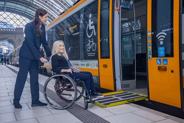 Auf dem Bild ist eine Zugbegleiterin, die einer Person im Rollstuhl beim Einstieg in den Trilex - Zug assistiert.