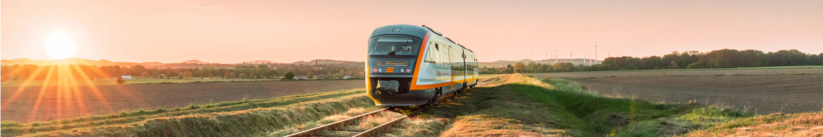 Ein trilex Zug fährt bei Sonnenaufgang über eine eingleisige Strecke durch eine offene Landschaft. Die Sonne steht tief am Horizont und taucht die Felder in warmes Licht.