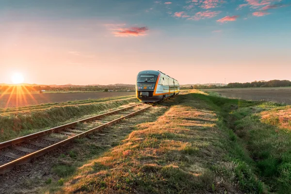 Ein trilex Zug fährt bei Sonnenaufgang über eine eingleisige Strecke durch eine offene Landschaft. Die Sonne steht tief am Horizont und taucht die Felder in warmes Licht.