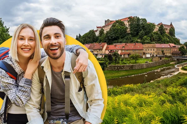 Paar in Wanderkleidung steht im Vordergrund. Im Hintergrund ist eine Landschaft mit historischen Gebäuden zu sehen.