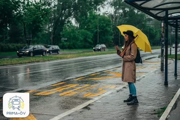 Frau steht an einem verregneten Tag mit einem gelben Regenschirm an einer Bushaltestelle und wartet.