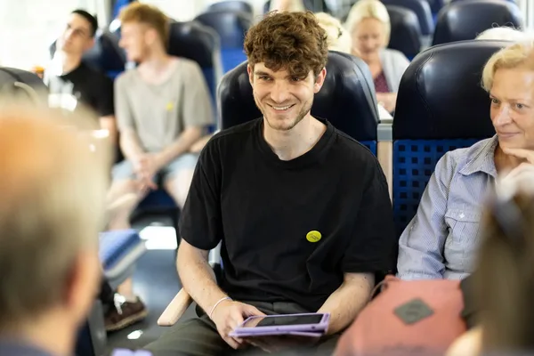 Eine Gruppe von Reisenden sitzt gemeinsam in einem Zug. Der Fokus liegt auf einem Mann, der mit Menschen ins Gespräch kommt.