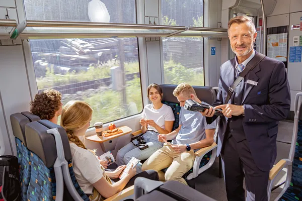 Das Bild zeigt eine Person in Uniform, die ein mobiles Gerät für den Ticketverkauf im trilex-Zug verwendet. Im Hintergrund sind große Fenster sowie eine Reisegruppe