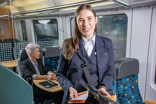 Das Bild zeigt eine Person in Uniform, die ein mobiles Gerät für den Ticketverkauf sowie einen Fahrplan in den Händen hält. Im Hintergrund ist eine Sitzecke mit einer Reisenden.