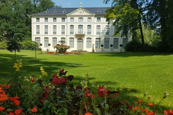 Das Bild zeigt das Sommerpalais im Greizer Park, ein großes, weißes, zweistöckiges Gebäude mit zahlreichen Fenstern und einem zentralen Balkon über dem Haupteingang. Das Gebäude hat ein symmetrisches Design mit dekorativen Elementen an der Fassade, einschließlich eines dreieckigen Giebels im oberen Zentrum. Vor dem Palais befindet sich ein gepflegter Rasen mit bunten Blumen im Vordergrund und Bäumen, die das Gebiet umgeben.