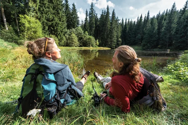 Das Bild zeigt zwei Personen, die auf dem Gras nahe einem kleinen See oder Teich im Hüttenbachtal sitzen, umgeben von einem dichten Wald. Die Person links trägt eine blaue Jacke und hat einen grün-schwarzen Rucksack, während die Person rechts ein rotes Shirt trägt und einen grauen Rucksack hat. Beide blicken auf das Wasser, mit Bäumen im Hintergrund unter einem teilweise bewölkten Himmel.