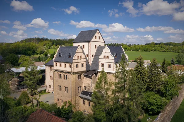 Das Bild zeigt einen großen, historischen Gebäudekomplex, umgeben von üppigem Grün und Bäumen. Die Struktur scheint ein Kloster oder eine Burg zu sein, mit mehreren Ebenen, steilen Dächern und zahlreichen Fenstern. Der Himmel ist klar mit vereinzelten Wolken, und die umliegende Landschaft umfasst Felder und weitere Gebäude in der Ferne.