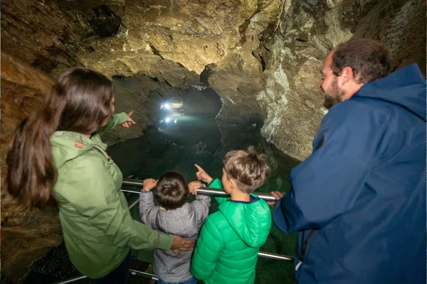 Das Bild zeigt vier Personen, zwei Erwachsene und zwei Kinder, die auf einem Metallgeländer in einer Höhle stehen. Die Erwachsenen tragen Jacken, und die Kinder sind ebenfalls warm angezogen. Die Höhle hat felsige Wände und ist schwach beleuchtet mit künstlichem Licht. Einer der Erwachsenen zeigt auf etwas in der Höhle, während eines der Kinder in die gleiche Richtung zeigt.