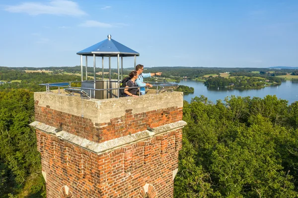 Das Bild zeigt einen Backstein-Aussichtsturm mit einem Metalldach und einem Geländer oben. Zwei Personen stehen auf der Aussichtsplattform und blicken auf eine malerische Landschaft mit dichten grünen Wäldern und einem großen Gewässer im Hintergrund. Der Himmel ist klar mit einigen verstreuten Wolken.
