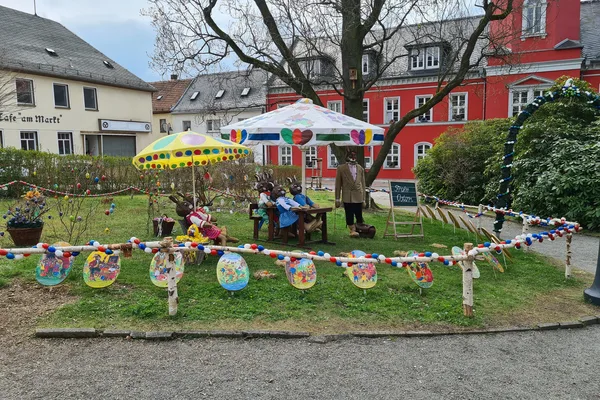 Auf einer Wiese, die sich auf dem Rathausvorplatz befindet, sitzen große Hasenfiguren mit Kleidung auf Schulbänken, die mit Sonnenschirmen überdacht sind. Die Wiese ist mit einer osterlichen Girlande umzäunt.