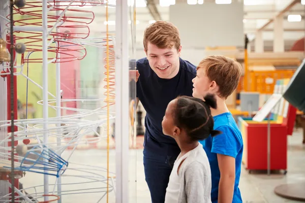 Das Bild zeigt einen Jungen und ein Mädchen, die gemeinsam mit einem jungen Mann auf ein Ausstellungsstück in einem wissenschaftlichen Museum schauen.