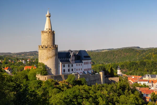 Das Bild zeigt die Osterburg in Weida, die mit einem großen Turm oberhalb der Stadt auf einem Hügel thront. Sie ist umgeben von dichtem Wald und auch im Hintergrund sind hügelige Wälder zu sehen.