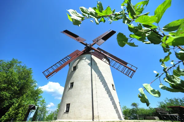 Das Bild zeigt eine alte Windmühle von unten. Sie ist in hellem grau und hat rostfarbene Rotorblätter.