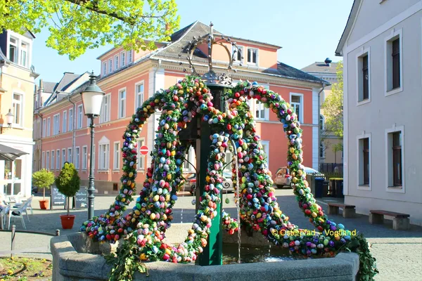 Das Bild zeigt eine hohe Krone aus Ästen, die mit Ostereiern geschmückt ist. Sie ist auf einem Brunnen auf einem Marktplatz platziert.