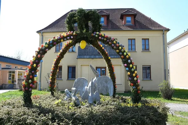 Das Bild zeigt eine hohe Krone aus Ästen, die mit Ostereiern geschmückt ist. Sie umspannt Sträucher, in deren Mitte mehrere Vogelstatuen abgebildet sind.