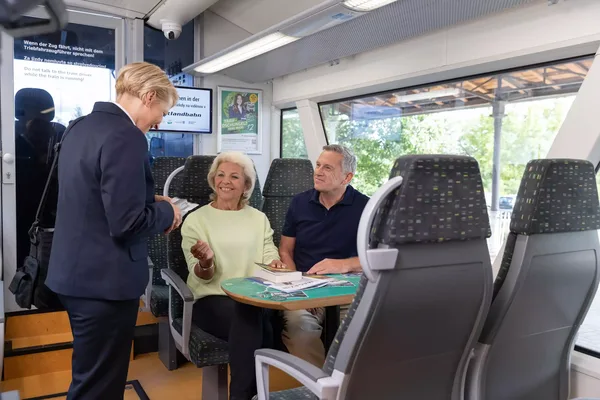 Das Bild zeigt eine Person in Uniform, die ein älteres Paar in der Vogtlandbahn über die Ticketpreise berät und einen Fahrschein kontrolliert.