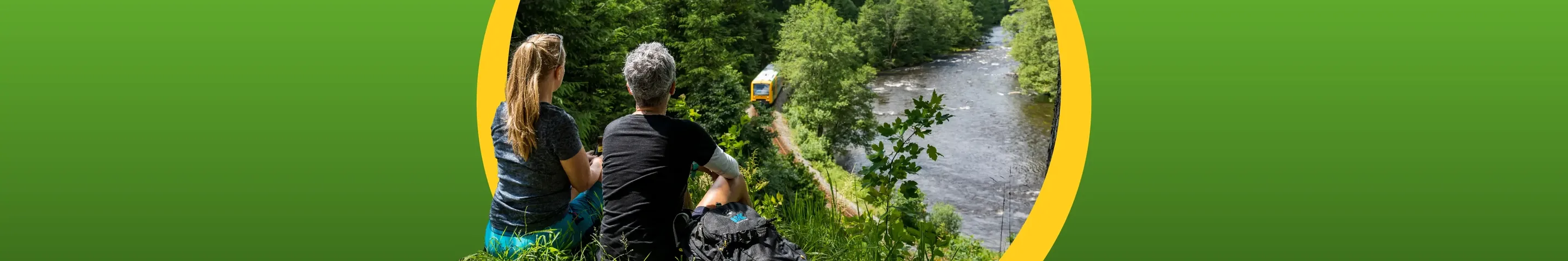 Zwei Personen blicken von einem Berg aus auf die waldbahn herunter, die an einem Fluss entlang fährt.