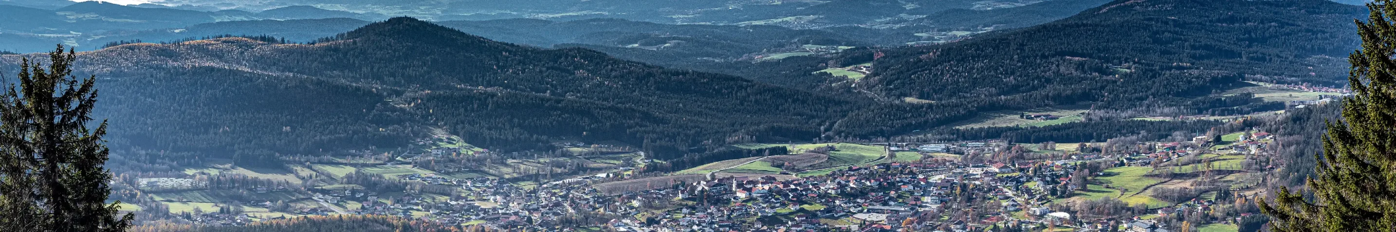 Blick von oben, auf eine Ortschaft, die von vielen Hügeln und Bergen voller Bäume umgeben ist.