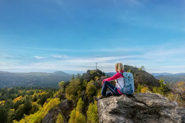 Eine Frau sitzt auf deinem Felsbrocken und blickt auf die Hügel, Berge und Wälder des Bayerischen Walds, die sie vom Silberberg aus sieht.