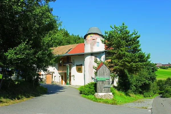 Die Gläserne Scheune in Viechtach mit einem kunstvoll gestalteten Turm, der im oberen Teil ein Gesicht eines Mannes zeigt und das Dach als Hut inszeniert.