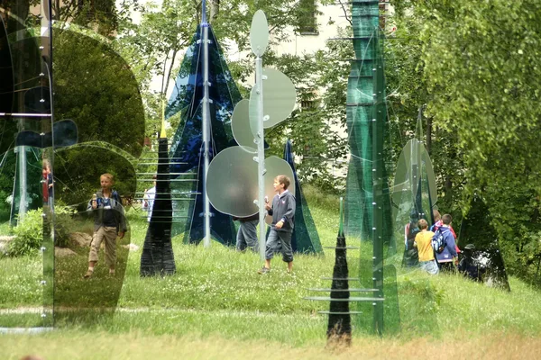 Menschen schlendern durch Kunstinstallationen aus verschiedenfarbigem Glas in einem Waldstück.
