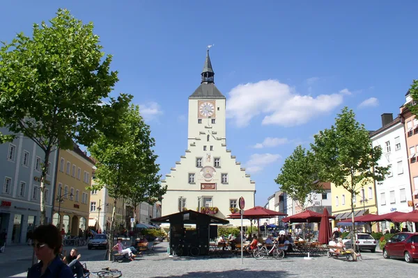 Ein großer Platz mit vielen Sonnenschirmen, Sitzgelegenheiten und einem großen Gebäude mit Turm.