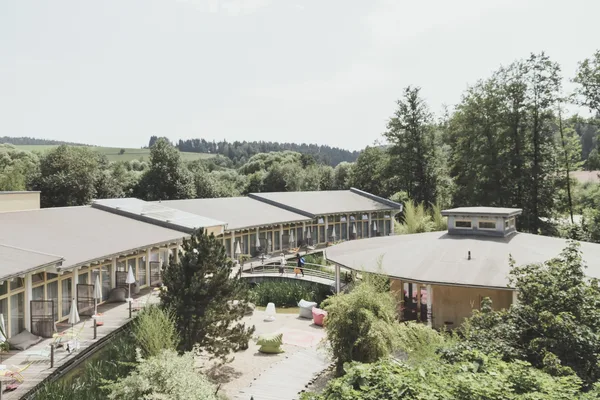 Blick von oben auf einen Gebäudekomplex aus einem kleinen runden und einem langgezogenen Gebäude, der durch eine kleine Brücke über einen Bach verbunden wird. Im Hintergrund sind Bäume und Wälder zu sehen.