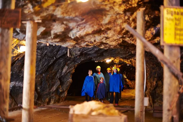 Eine Gruppe aus Erwachsenen und Kindern mit Schutzhelmen und blauen Kitteln geht durch einen niedrigen Tunnel innerhalb eines Bergwerks.