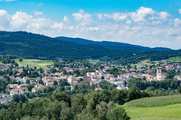 Blick auf eine Stadt umgeben von Wiesen und bewaldeten Hügeln und Bergen.