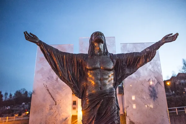 Eine goldfarbene Jesusstatue mit ausgebreiteten Armen vor drei Milchglasscheiben.