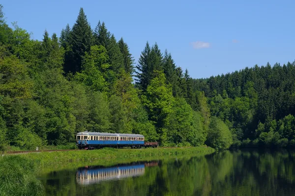 Die blau-weiße Wanderbahn fährt an einem Fluss entlang neben einem Waldstück.