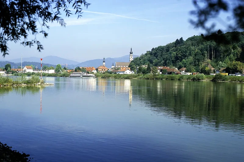 Blick auf die breite und ruhige Donau, im Hintergrund ist eine Stadt mit Kirchtürmen zu sehen.