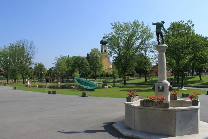 Ein Brunnen mit einer Statue in einem weitläufigen Park. Im Hintergrund Glasinstallationen und Kunstwerke auf der Parkwiese mit vielen Bäumen.