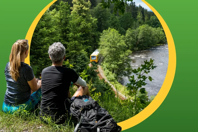 Zwei Personen blicken von einem Berg aus auf die waldbahn herunter, die an einem Fluss entlang fährt.