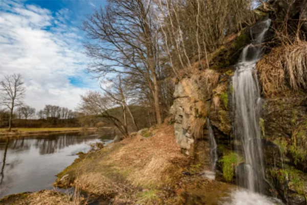 Ein Wasserfall, der aus einem Waldstück heraus, Felsen hinunter führt und links davon ein ruhiges Gewässer, in das der Wasserfall mündet.