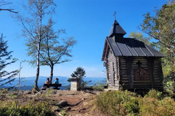 Eine Kapelle vor blauem Himmel auf einem Berggipfel mit einer Sitzbank und Ausblick über den Bayerischen Wald.