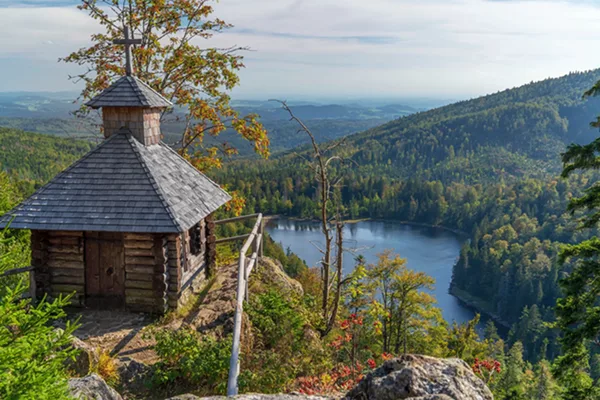 Blick auf eine Kapelle auf einem Berggipfel mit Blick auf ein ruhiges Gewässer und dichten Wald auf Hügeln und Bergen.