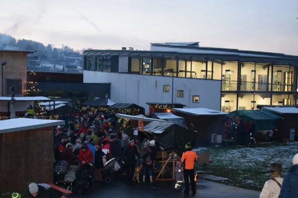 Ein vollbesuchter Weihnachtsmarkt mit einzelnen Buden neben einem Gebäude mit großen Fenstern.